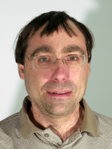 Herr Gould mit Brille, braunem Pulli und hellem Kragen, direkter Blick.